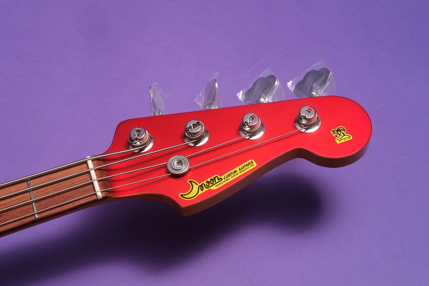 Close-up of a red electric bass guitar headstock with tuning pegs on a purple background